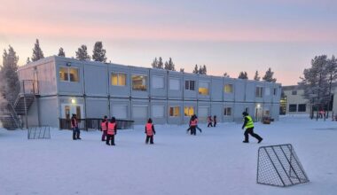 Barn i fargerike vester leker ute i snøen ved midlertidige bygninger. Noen sitter på bakken, mens andre står og beveger seg, med et fotballmål i forgrunnen. Omgivelsene er preget av snødekte områder og trær i bakgrunnen. Det er klart vær, og himmelen har et svakt fargespill fra soloppgang. (Bildebeskrivelsen er laget av en KI-tjeneste)