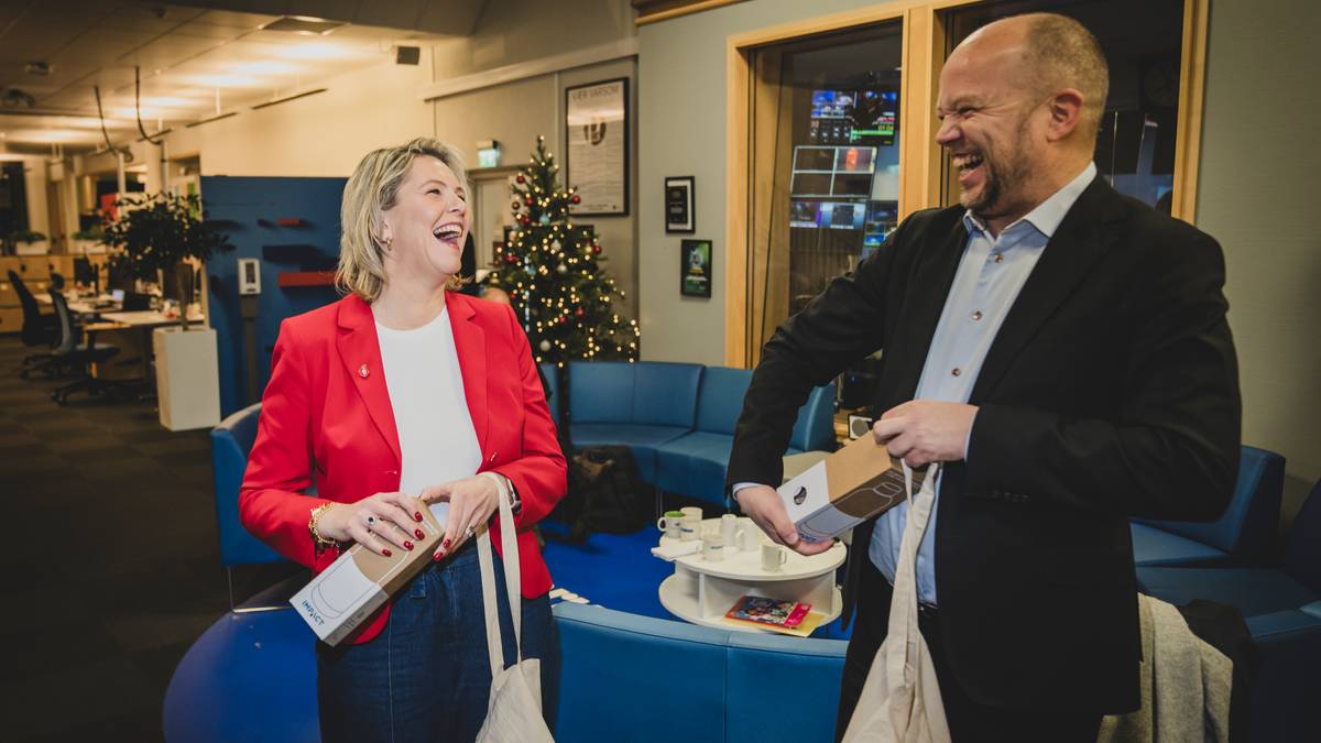 To personer står sammen i et moderne kontormiljø, hvor julepynt er synlig i bakgrunnen. Den ene personen, iført en rød blazer, holder en pakke og en tote-bag, mens den andre, med skjorte og dressjakke, også bærer en eske. Det er møbler i blått og lys grønne toner i rommet, og det er en kaffekopp på et bord i nærheten. Kontoret ser ut til å være i aktivitet med flere synlige arbeidsplasser. (Bildebeskrivelsen er laget av en KI-tjeneste)