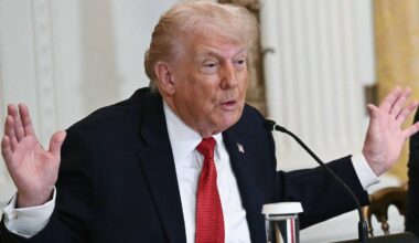 En mann sitter ved et bord, han har på seg en mørk dress med en rød slips. Han gestikulerer med begge hender mens han snakker. Det er en mikrofon plassert på bordet foran ham, og det er en annen person delvis synlig til høyre i bildet. Omgivelsene ser ut som en offisiell setting med dekorative elementer i bakgrunnen. (Bildebeskrivelsen er laget av en KI-tjeneste)