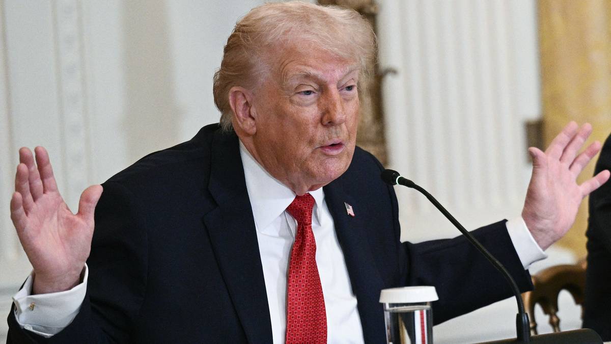 En mann sitter ved et bord, han har på seg en mørk dress med en rød slips. Han gestikulerer med begge hender mens han snakker. Det er en mikrofon plassert på bordet foran ham, og det er en annen person delvis synlig til høyre i bildet. Omgivelsene ser ut som en offisiell setting med dekorative elementer i bakgrunnen. (Bildebeskrivelsen er laget av en KI-tjeneste)