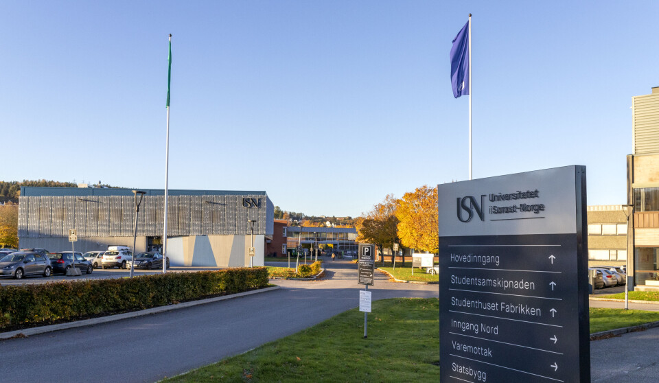 Veiskilt ved et moderne campusområde med bygninger, gress og flagg i bakgrunnen.