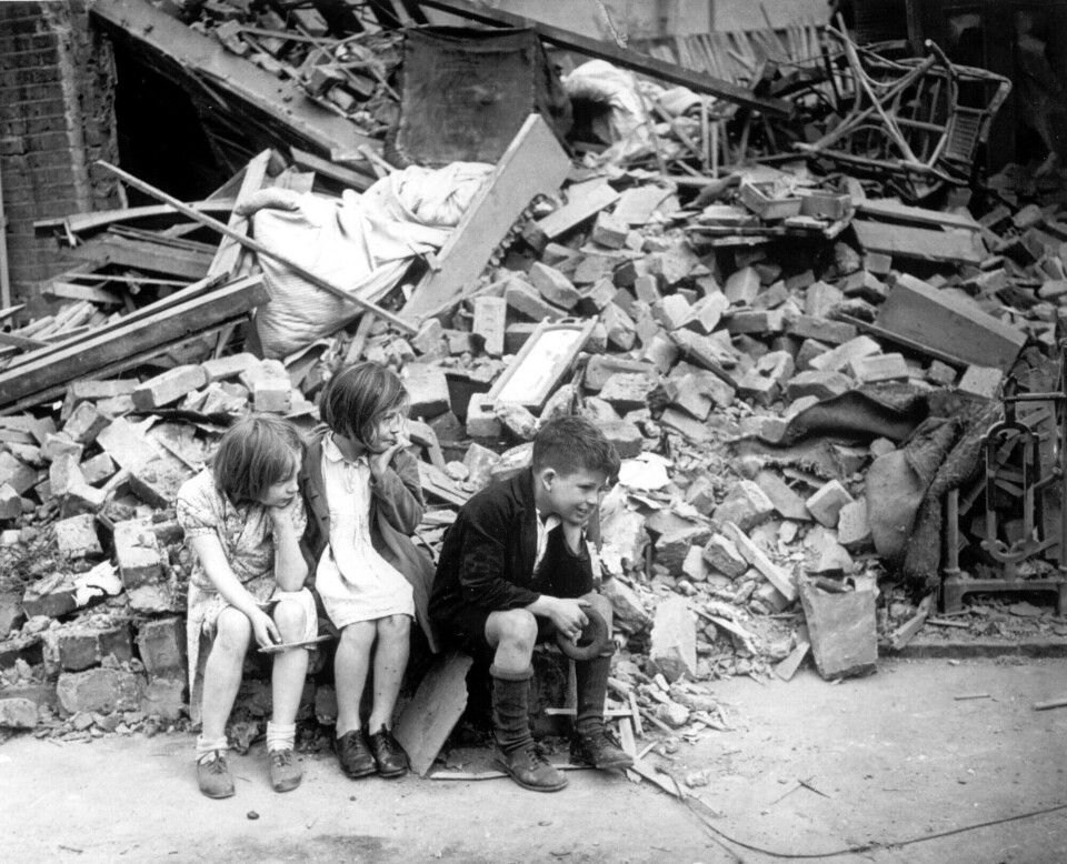 Tre barn sitter foran en stor haug med murstein og ruiner i Øst-London i 1940.
