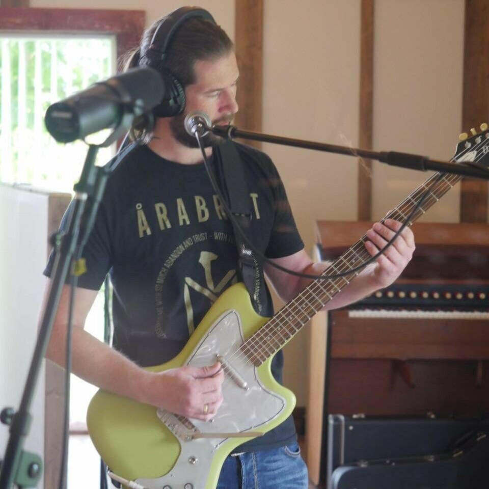 Mann med hodetelefoner spiller elektrisk gitar og synger i mikrofon i et rom med piano.