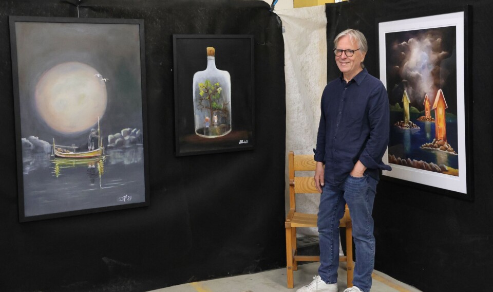 En mann står ved siden av tre innrammede malerier utstilt på svarte vegger i et gallerirom.