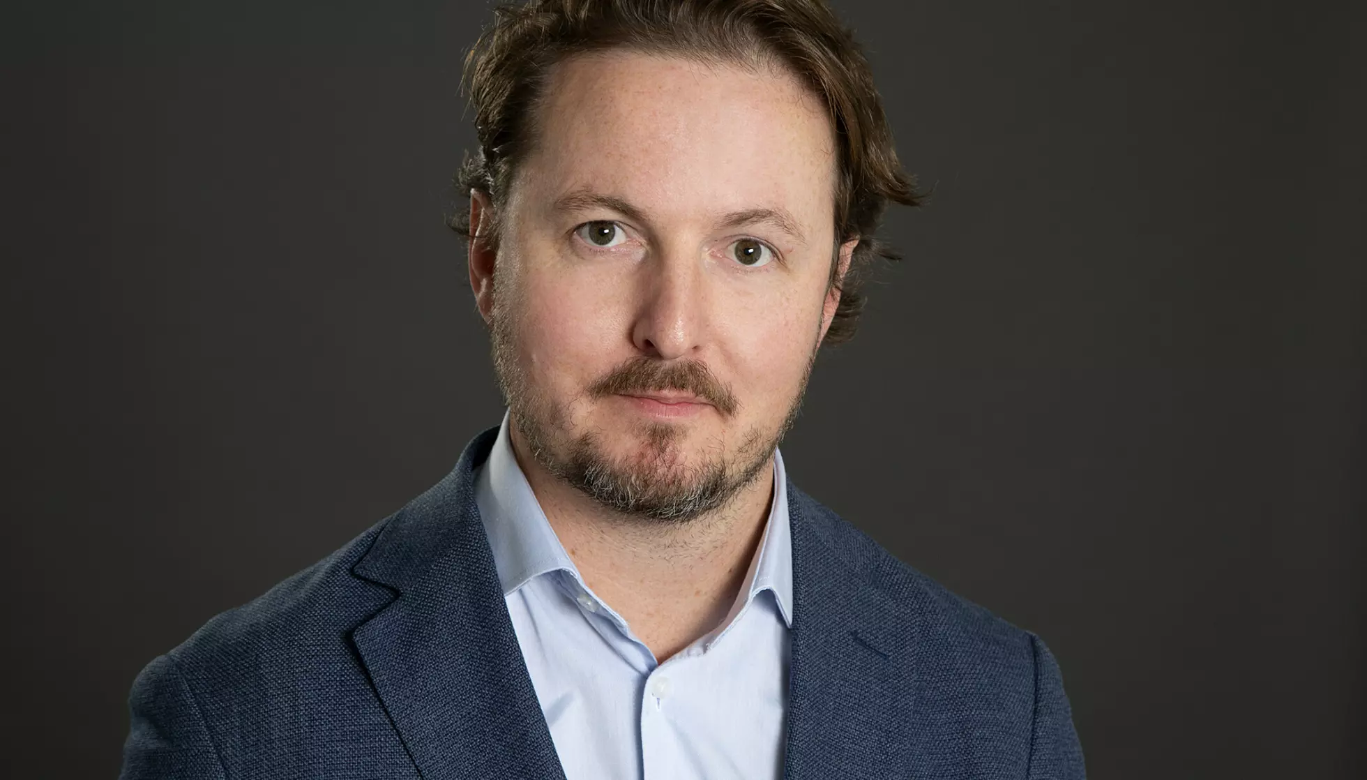 Olav Brekke Mathisen i et mørlt rom. ikledd blå dress. brunt hår til siden og litt skjegg. Nøytralt ansikt
