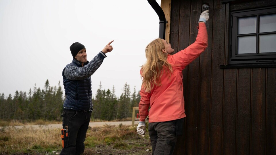 To personer ved en mørk trehytte, en peker opp og en rekker mot veggen.