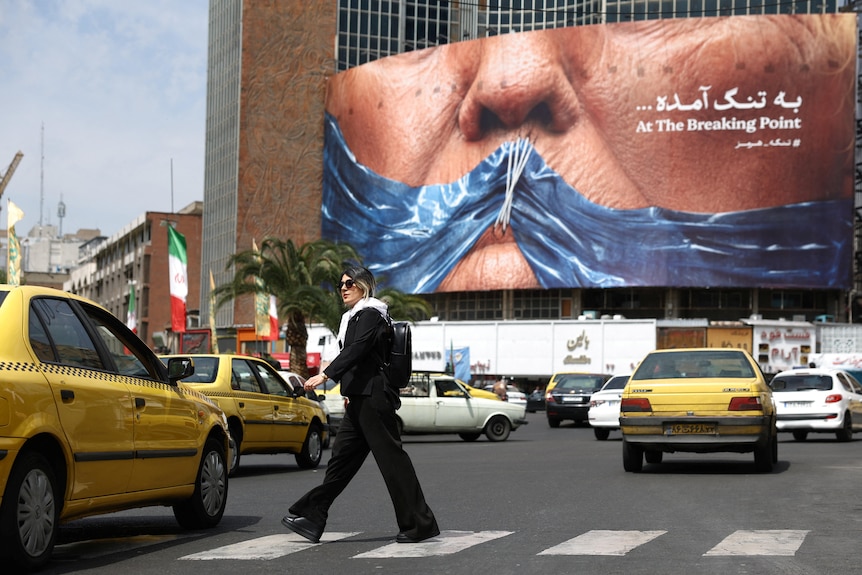 A woman walks across a Zebra crossing near some Taxis. In the background, a billboard depicts Trump with his mouth sewn shut.