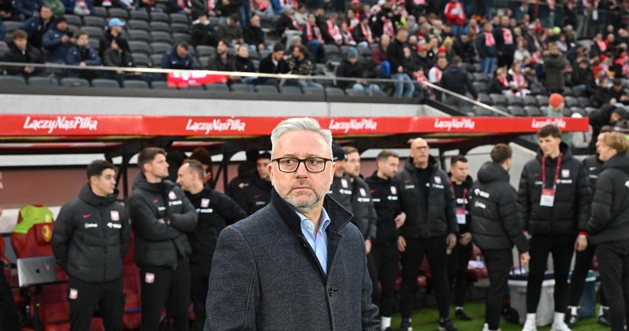 Mężczyzna w okularach i koszuli z krótkim rękawem na trybunach stadionu, w tle rozmyci kibice i elementy stadionu sportowego.