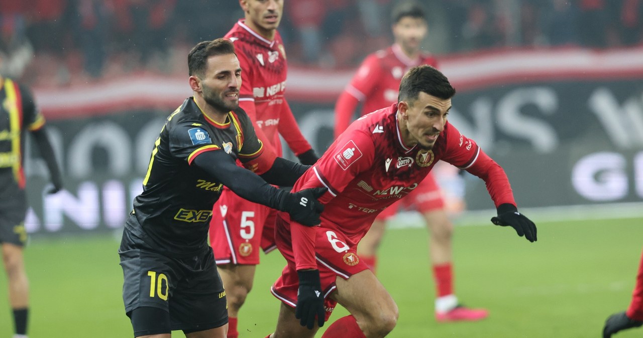 Bramkarz ubrany w niebieski strój łapie piłkę podczas meczu piłki nożnej, na trybunach widać rozmytą publiczność.