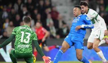 Piłkarz w białej koszulce z rozłożonymi rękami świętuje zdobycie gola, wokół niego zespołowi koledzy w podobnych strojach wyrażają radość, w tle widać trybuny pełne kibiców.