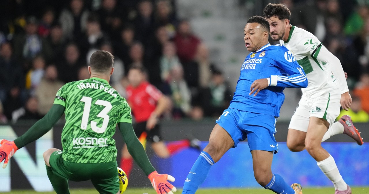 Piłkarz w białej koszulce z rozłożonymi rękami świętuje zdobycie gola, wokół niego zespołowi koledzy w podobnych strojach wyrażają radość, w tle widać trybuny pełne kibiców.