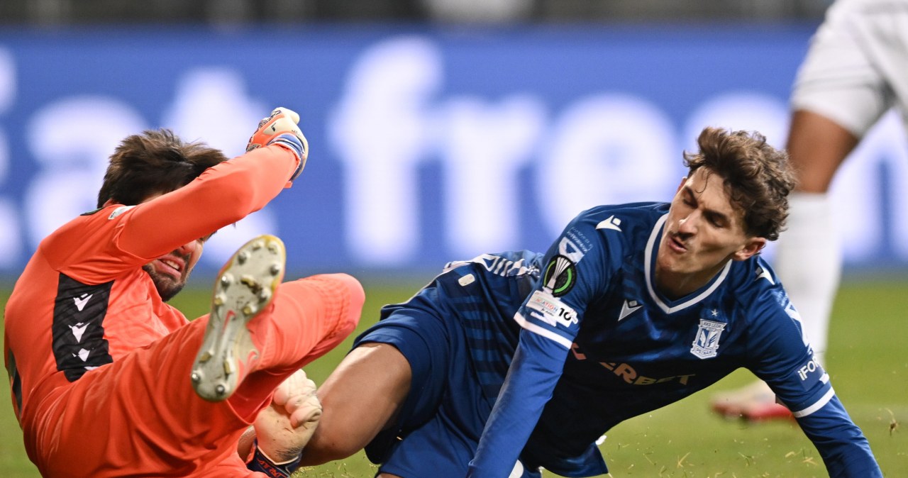 W akcji sportowej kilku piłkarzy na murawie stadionu, zawodnik w niebieskim stroju prowadzi piłkę, ścigany przez zawodników w białych strojach z logo INEOS, w tle rozmazana sylwetka bramkarza.