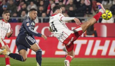 Piłkarz w ciemnym stroju leżący na murawie z nogami uniesionymi w powietrzu podczas upadku, w tle sylwetki zawodników w biało-czerwonych strojach oraz rozmyte elementy stadionu.