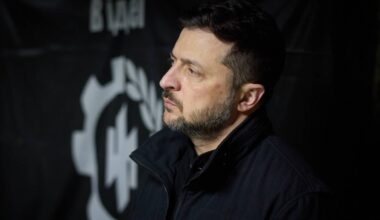 Wołodymyr Zełenski, prezydent Ukrainy, w profilu, z poważnym wyrazem twarzy, na tle czarnej flagi z symbolem