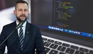 Minister obrony narodowej Władysław Kosiniak-Kamysz przemawia przed mikrofonem, z laptopem i kodem programu komputerowego w tle.