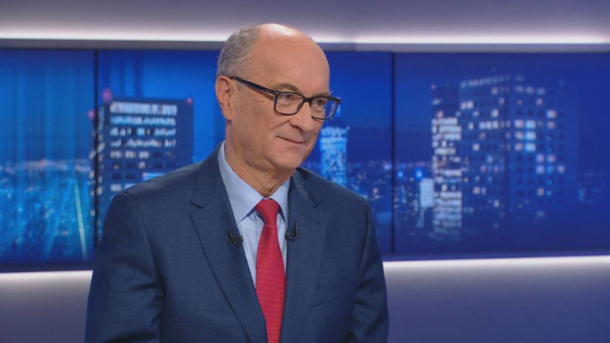 Mężczyzna w garniturze i czerwonym krawacie z okularami na nosie, siedzący na tle niebieskich ekranów z widokiem nocnego miasta.