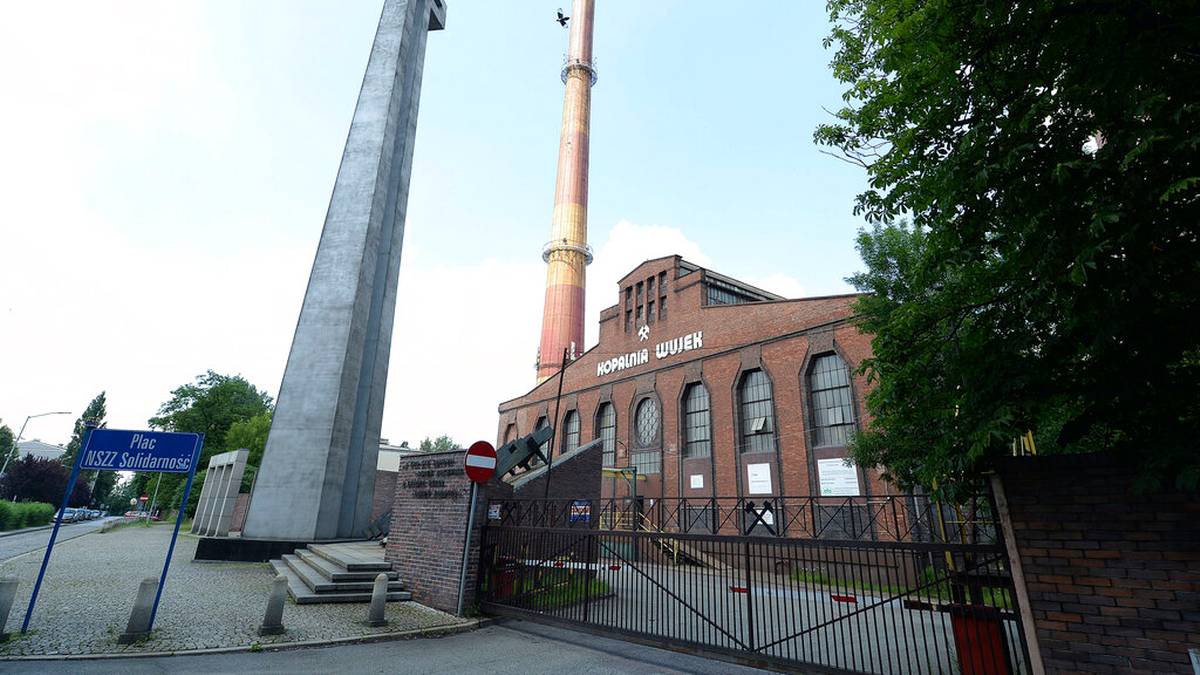 Monumentalny monument krzyża obok ceglanego budynku przemysłowego z kominem w Katowicach.