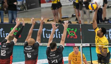 Mecz siatkówki, gracz w żółtym stroju atakuje, czterech graczy w czarnych strojach próbuje zablokować.