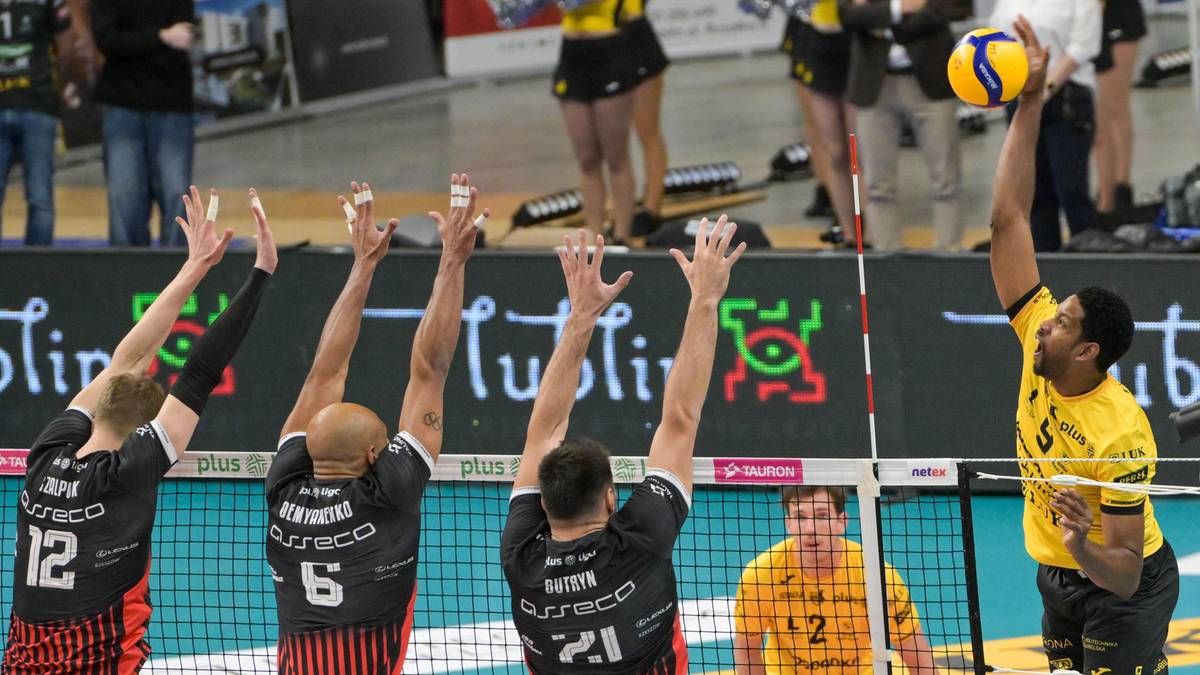 Mecz siatkówki, gracz w żółtym stroju atakuje, czterech graczy w czarnych strojach próbuje zablokować.