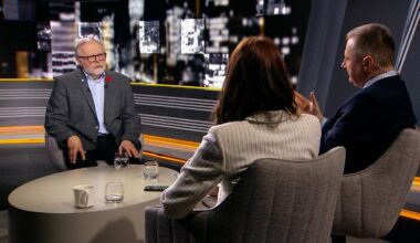 Profesor Szymon Malinowski siedzi przy stole w studiu telewizyjnym, rozmawiając z kobietą i mężczyzną. Na stole stoją szklanki i filiżanki.