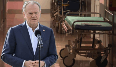 Premier Donald Tusk przemawia na tle szpitalnego korytarza z wózkami medycznymi.