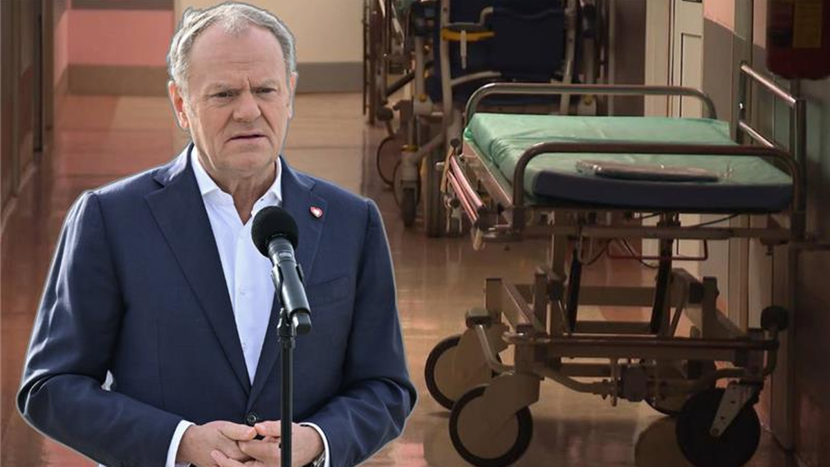 Premier Donald Tusk przemawia na tle szpitalnego korytarza z wózkami medycznymi.