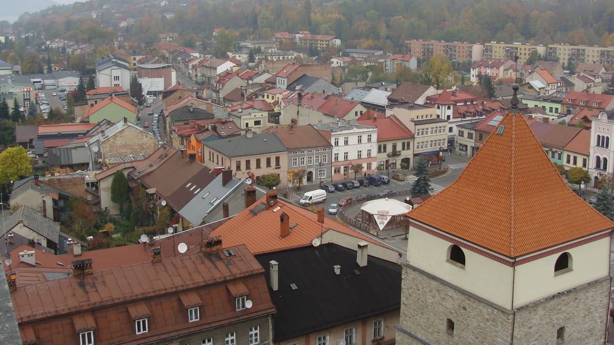 Widok panoramy miasta z widocznymi dachami budynków i wieżą obronną, w tle wzgórza, wszystko spowite mgłą lub smogiem.