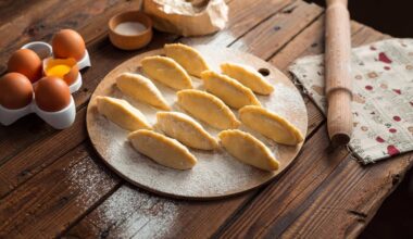 Surowe pierogi ułożone na desce, obok wałek, mąka i jajka.