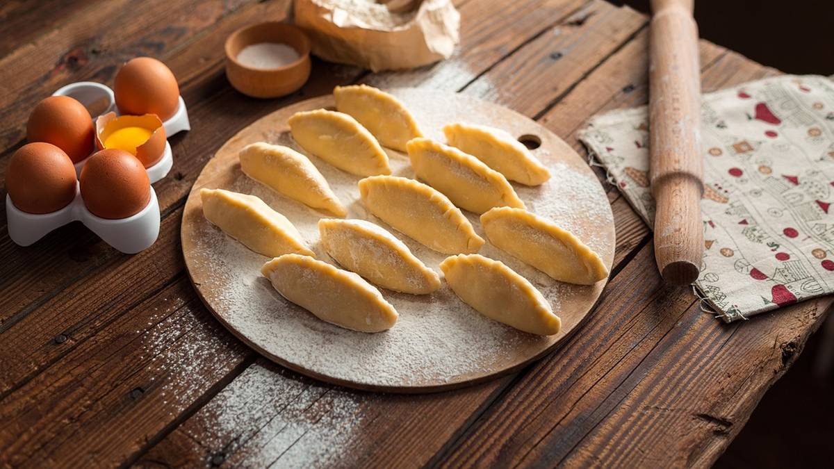 Surowe pierogi ułożone na desce, obok wałek, mąka i jajka.