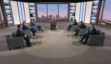 Grupa mężczyzn w garniturach siedzi w studio telewizyjnym, dyskutując przy okrągłym stoliku.