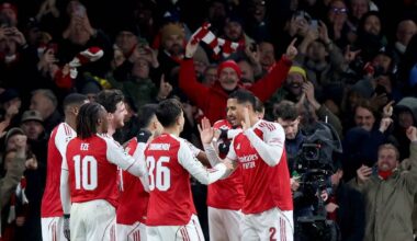 Grupa piłkarzy w czerwonych koszulkach z białymi numerami na plecach, ciesząca się po strzelonej bramce, podczas gdy kibice na trybunach w tle również wyrażają radość.