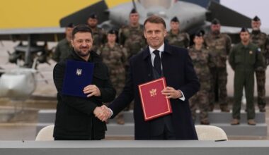 Prezydent Ukrainy Wołodymyr Zełenski i prezydent Francji Emmanuel Macron stoją obok siebie, uśmiechają się i ściskają dłonie. Zełenski trzyma niebieski dokument, a Macron czerwony. W tle widać myśliwiec Rafale i grupę żołnierzy.