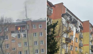 Uszkodzony blok mieszkalny po eksplozji, widoczne wyrwane ściany i balkony, dym unoszący się z jednego z mieszkań.