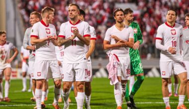 Piłkarze reprezentacji Polski w białych strojach stoją na boisku piłkarskim, w tle kibice.