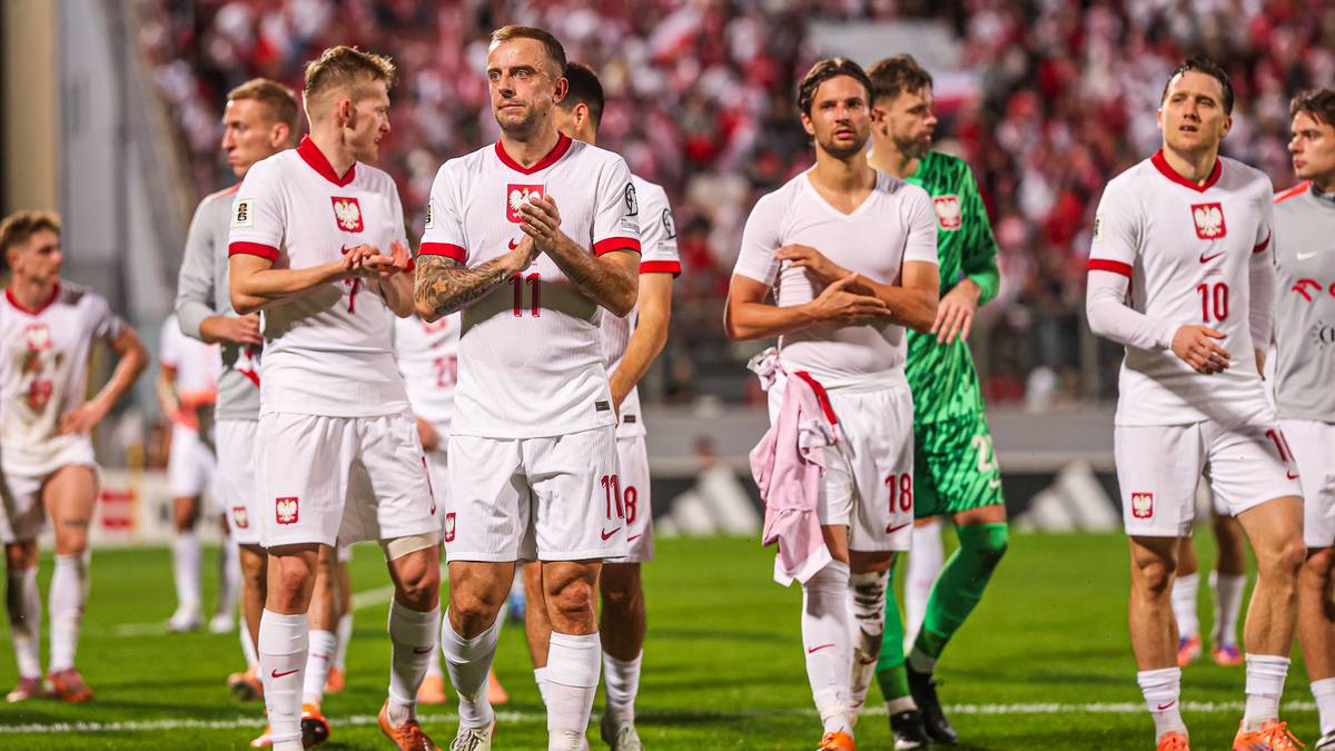 Piłkarze reprezentacji Polski w białych strojach stoją na boisku piłkarskim, w tle kibice.