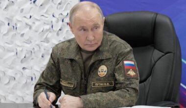 Władimir Putin w mundurze wojskowym siedzi przy biurku i pisze, na niebieskim tle z rosyjskimi napisami.