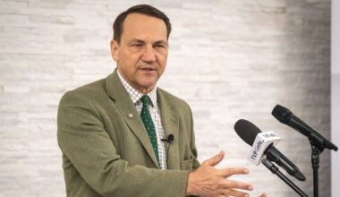 Radosław Sikorski przemawia przy mównicy z mikrofonami, w jasnobrązowej marynarce i zielonym krawacie.