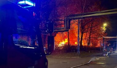 Nocny pożar na terenie zakładu przemysłowego, widoczne płomienie i wóz strażacki z wężem gaśniczym.