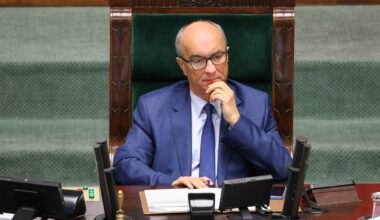 Mężczyzna w garniturze i okularach siedzi za stołem w sali sejmowej, opierając podbródek na dłoni.