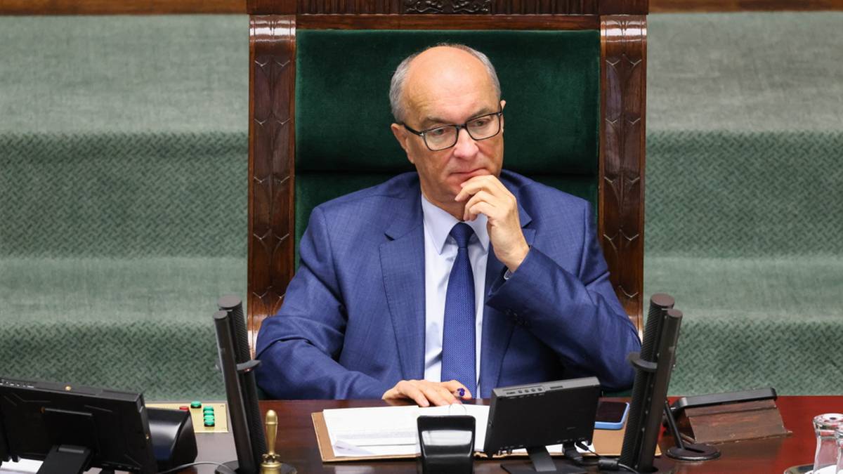 Mężczyzna w garniturze i okularach siedzi za stołem w sali sejmowej, opierając podbródek na dłoni.