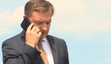 Mężczyzna w ciemnym garniturze i jasnej koszuli, z krawatem, rozmawia przez czarny smartfon trzymany przy uchu. Tło jest jasnoniebieskie.