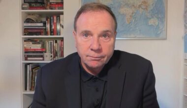 Generał Frederick Hodges w garniturze siedzi przed regałami z książkami, na ścianie wisi mapa świata.