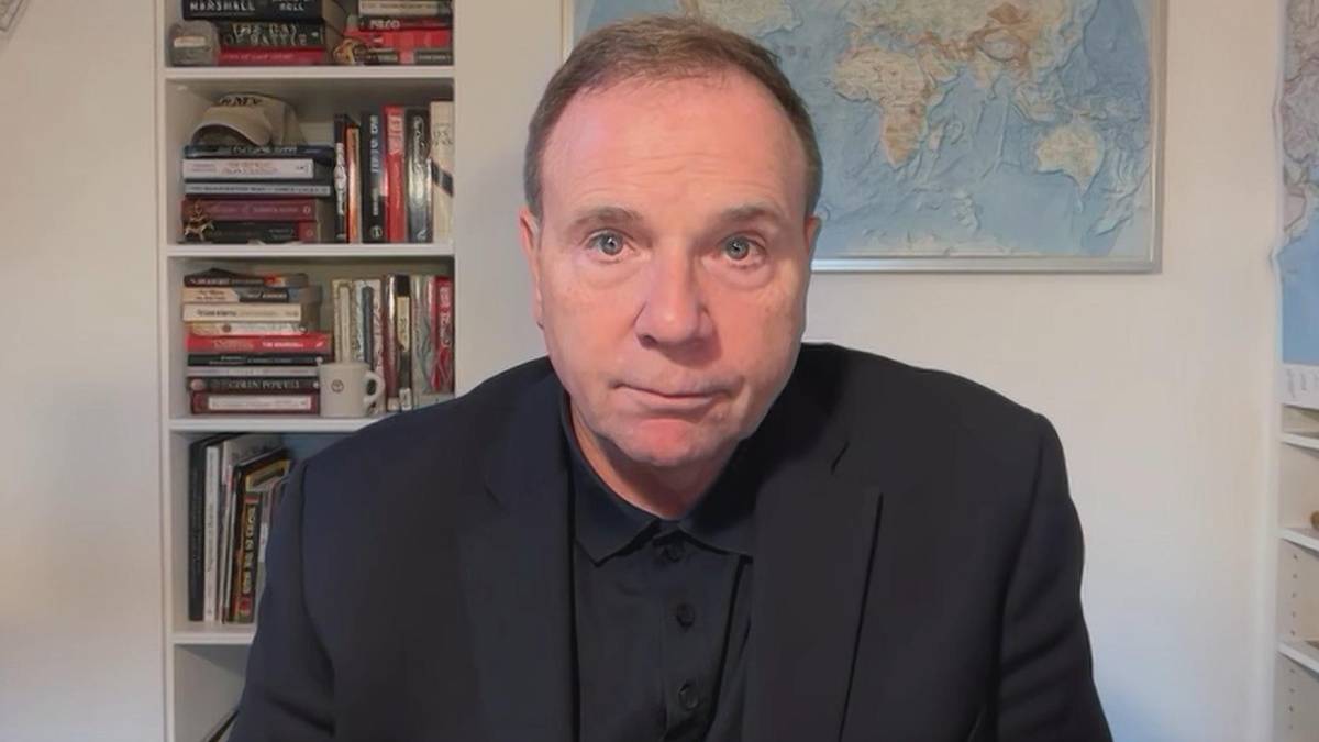 Generał Frederick Hodges w garniturze siedzi przed regałami z książkami, na ścianie wisi mapa świata.