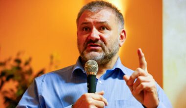 Mężczyzna w niebieskiej koszuli trzyma mikrofon i gestykuluje, przemawiając.