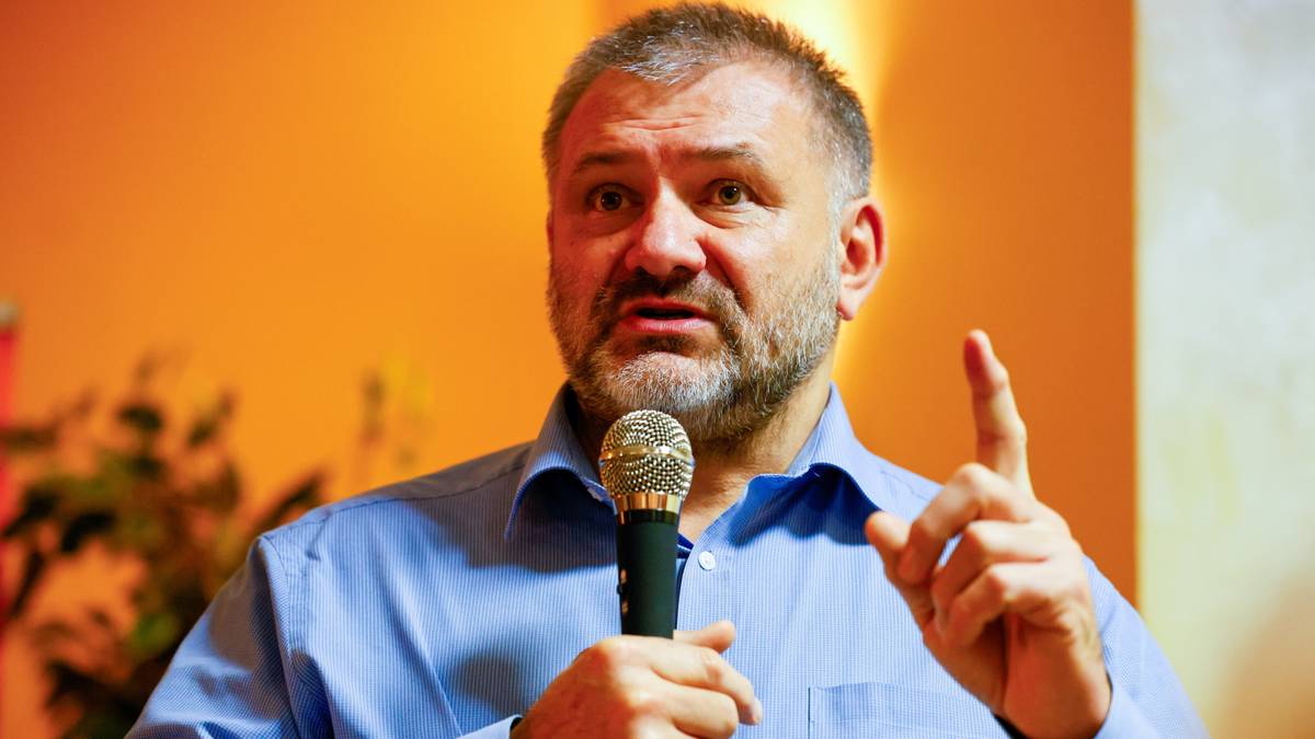 Mężczyzna w niebieskiej koszuli trzyma mikrofon i gestykuluje, przemawiając.