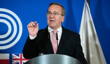 Boris Pistorius, niemiecki minister obrony, przemawia za mównicą na tle banera i flag państwowych.