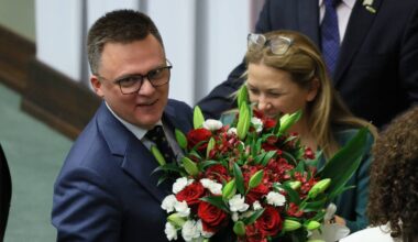 Szymon Hołownia w garniturze z okularami na nosie, uśmiechnięty, trzymający bukiet czerwonych i białych róż z zielonymi liśćmi.