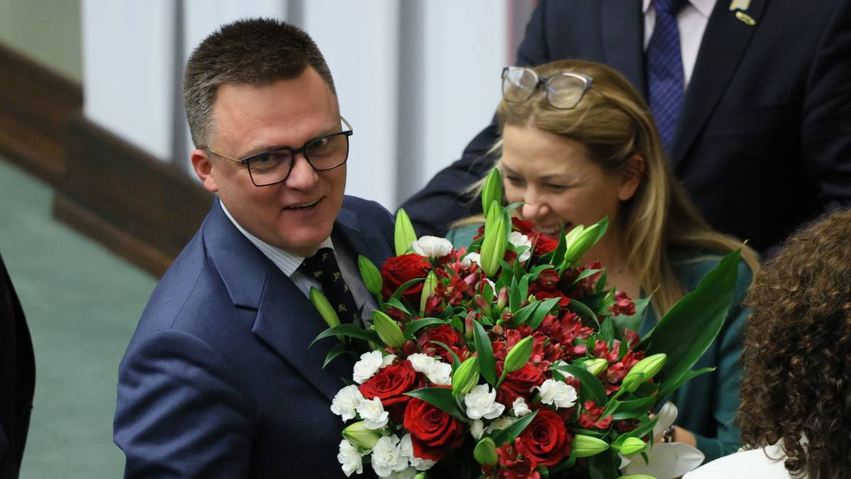 Szymon Hołownia w garniturze z okularami na nosie, uśmiechnięty, trzymający bukiet czerwonych i białych róż z zielonymi liśćmi.