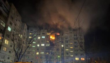 Pięciopiętrowy budynek mieszkalny w nocy, z którego wydobywa się ogień i dym. Na dole widoczne są wozy strażackie.