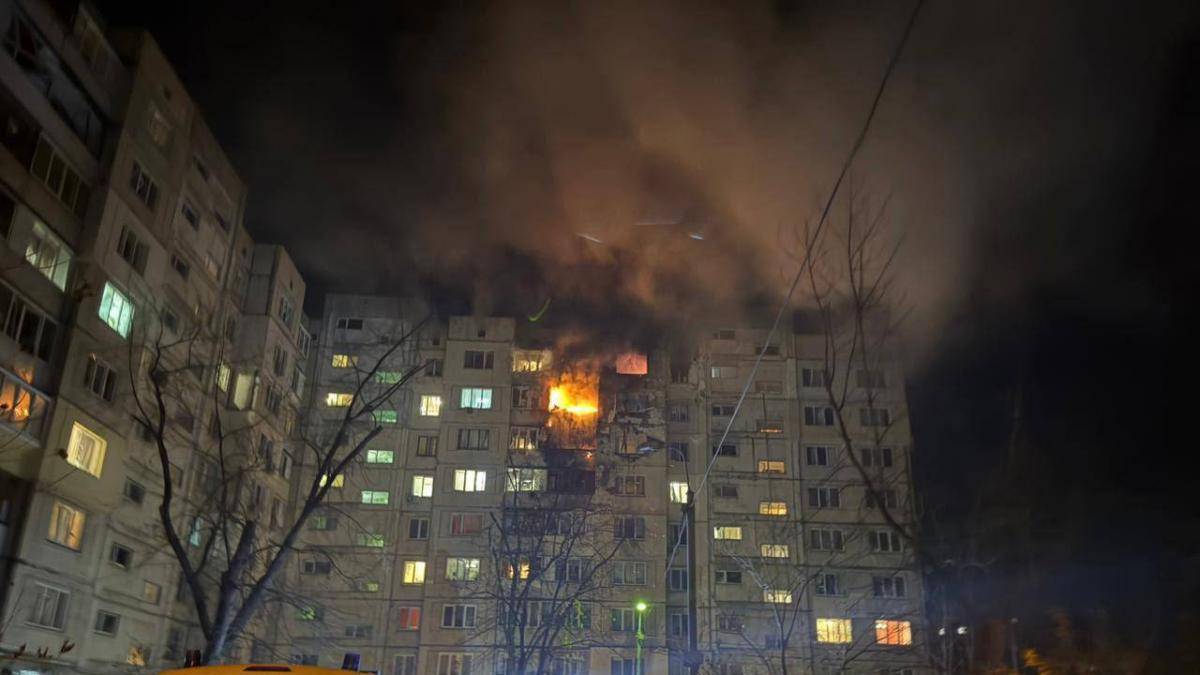 Pięciopiętrowy budynek mieszkalny w nocy, z którego wydobywa się ogień i dym. Na dole widoczne są wozy strażackie.